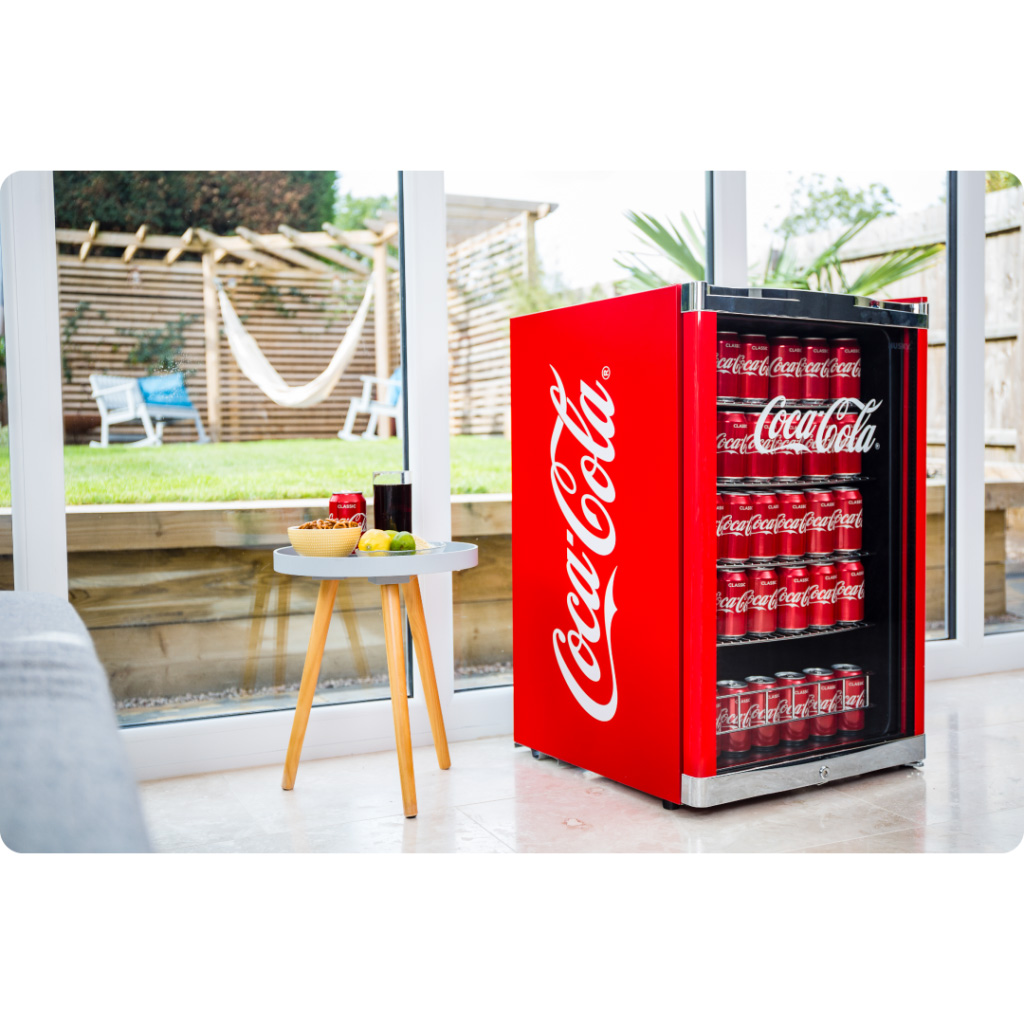 A Coca-Cola mini fridge is filled with Coca-Cola cans, standing beside a small table with drinks and snacks. The setting includes a glass-walled room overlooking a garden with a hammock.